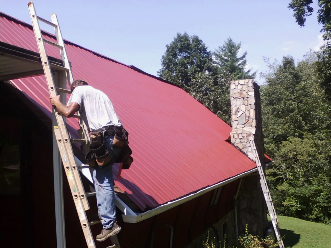 Roofer on a ladder beside a red metal roof and stone chimney — Soffit & Fascia Repair by Roofing Brothers in North Hampton